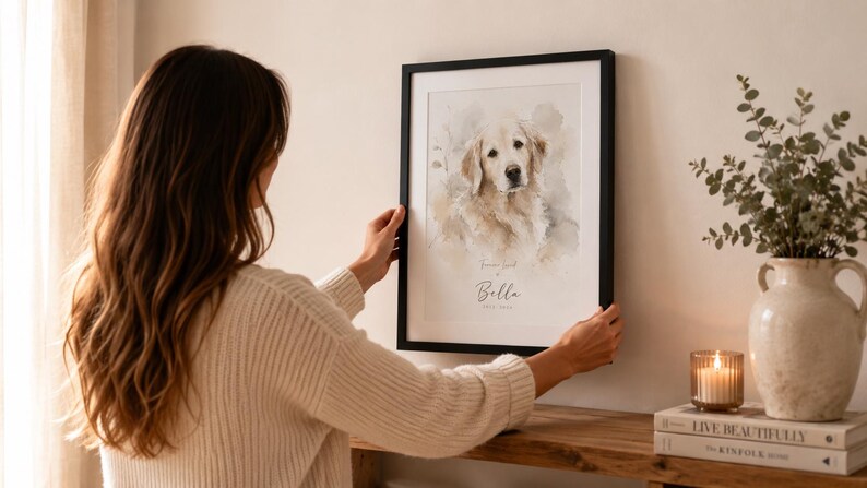 May include: A framed watercolor print of a Golden Retriever dog with the name "Bella" below the image. The print is in a black frame and is being held up by a person. The background includes a vase with greenery, a candle, and books.