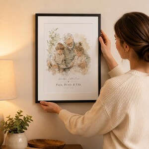 May include: A framed watercolor print featuring a grandfather with two grandchildren. The print has the text "Grandpa's Little Crew, Papa, Henry & Ella" and a date. The frame is black, and the print is being held by a person.