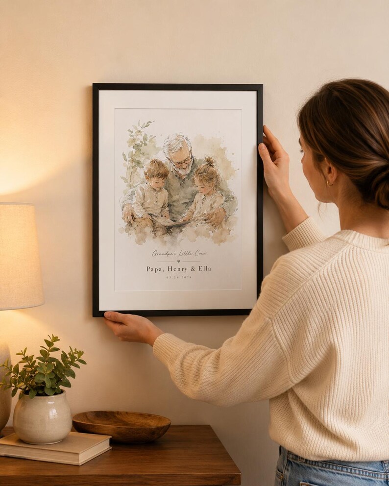 May include: A framed watercolor print featuring a grandfather with two grandchildren. The print has the text "Grandpa's Little Crew, Papa, Henry & Ella" and a date. The frame is black, and the print is being held by a person.