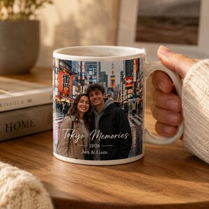 May include: White ceramic mug featuring a photograph of a couple in Tokyo, Japan. The mug has the text "Tokyo Memories 2026 Ava & Liam" printed on it. The mug is held by a person.