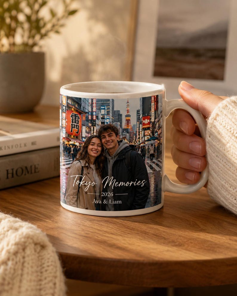 May include: White ceramic mug featuring a photograph of a couple in Tokyo, Japan. The mug has the text "Tokyo Memories 2026 Ava & Liam" printed on it. The mug is held by a person.