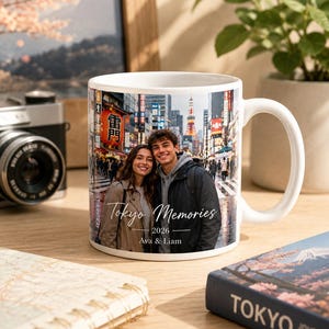 May include: White ceramic mug featuring a photograph of a couple in Tokyo, Japan, with the text "Tokyo Memories 2026 Ava & Liam." The mug has a large handle and is set on a wooden surface.