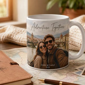 Pu&ograve; includere: Tazza in ceramica bianca con una foto di una coppia e la scritta "Adventure Together". La tazza riporta i nomi "Emily & Carter 2026". Un diario di viaggio e una macchina fotografica sono sullo sfondo.