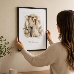 May include: A framed watercolor print featuring a couple embracing in front of a building. The print has a black frame and includes the text "Love Lives Here" and "The Wilsons Est. 2022". The print is being held by a person.