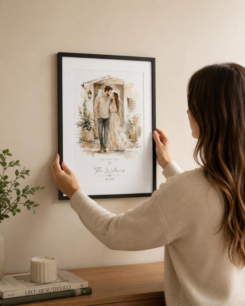 May include: A framed watercolor print featuring a couple embracing in front of a building. The print has a black frame and includes the text "Love Lives Here" and "The Wilsons Est. 2022". The print is being held by a person.