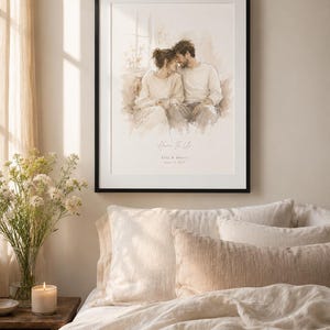 May include: A framed watercolor print featuring a couple embracing, with the text "Home Is Us" and a date. The artwork is in a black frame, displayed above a bed with cream-colored bedding and pillows. A lit candle and flowers are on a wooden nightstand.