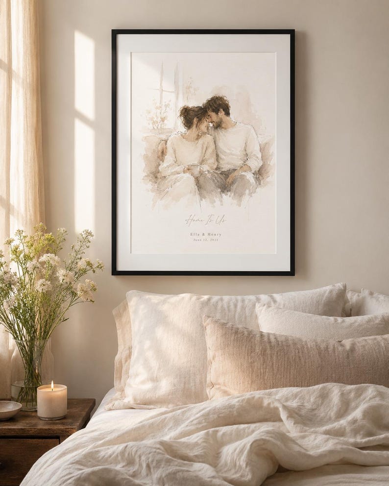 May include: A framed watercolor print featuring a couple embracing, with the text "Home Is Us" and a date. The artwork is in a black frame, displayed above a bed with cream-colored bedding and pillows. A lit candle and flowers are on a wooden nightstand.