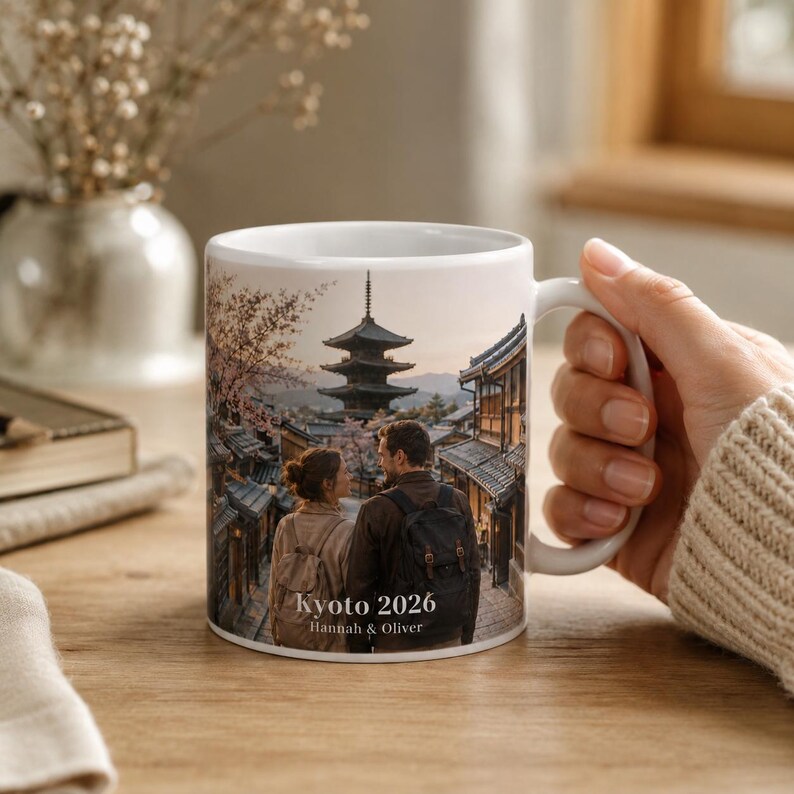 May include: White ceramic mug featuring a scenic image of Kyoto, Japan, with a pagoda and couple. The mug has the text "Kyoto 2026" and the names "Hannah & Oliver". The mug is held by a person.