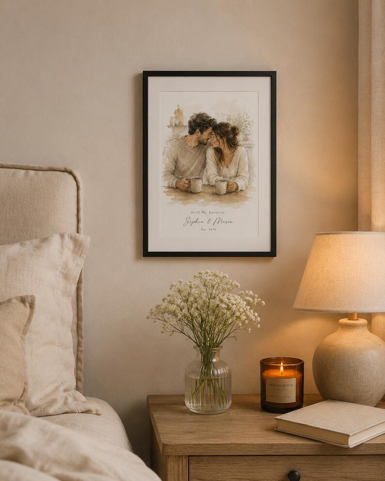 May include: A framed watercolor print depicts a couple sharing a tender moment, holding mugs. The print is in a black frame and hangs above a wooden nightstand. A vase of white flowers, a lit candle, and a lamp with a beige shade are also on the nightstand.