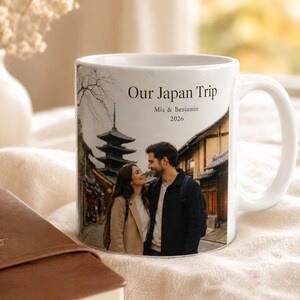 Pode incluir: Caneca de cer&acirc;mica branca com uma fotografia de um casal no Jap&atilde;o, com uma pagoda e edif&iacute;cios tradicionais. A caneca tem o texto "Our Japan Trip, Mia & Benjamin, 2026". Um di&aacute;rio de viagem castanho est&aacute; em primeiro plano.