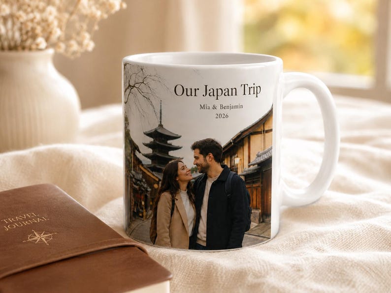 Pode incluir: Caneca de cer&acirc;mica branca com uma fotografia de um casal no Jap&atilde;o, com uma pagoda e edif&iacute;cios tradicionais. A caneca tem o texto "Our Japan Trip, Mia & Benjamin, 2026". Um di&aacute;rio de viagem castanho est&aacute; em primeiro plano.