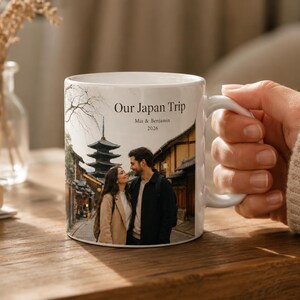 Pode incluir: Caneca de cer&acirc;mica branca com uma fotografia de um casal no Jap&atilde;o e o texto "Our Japan Trip". A caneca tem uma al&ccedil;a branca. O fundo inclui um vaso com flores secas e uma mesa de madeira. A caneca &eacute; segurada por uma pessoa.