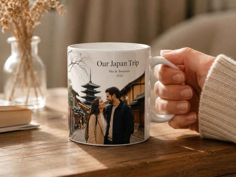 Pode incluir: Caneca de cer&acirc;mica branca com uma fotografia de um casal no Jap&atilde;o e o texto "Our Japan Trip". A caneca tem uma al&ccedil;a branca. O fundo inclui um vaso com flores secas e uma mesa de madeira. A caneca &eacute; segurada por uma pessoa.