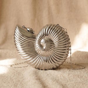 Peut inclure: Une pochette métallique argentée en forme de coquille de nautile. Le sac présente un extérieur nervuré en spirale avec une finition polie et comprend une fine bandoulière en chaîne argentée amovible fixée sur le dessus pour le transport.
