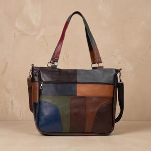May include: A multi-colored patchwork tote bag constructed from various leather panels in shades of brown, navy blue, olive green, tan, and dark gray. The bag features two shoulder straps, a horizontal zippered front pocket, and side hardware.
