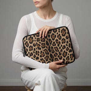 May include: A rectangular laptop sleeve featuring a leopard print pattern in shades of brown and black. The sleeve has a black zipper and trim. The product is held by a person wearing a white long-sleeved shirt and white pants.