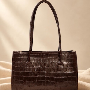 May include: A dark brown rectangular handbag featuring a textured crocodile skin pattern. The bag has two thin, matching vertical handles attached to the top edge with rectangular stitched tabs, providing a structured and uniform appearance.
