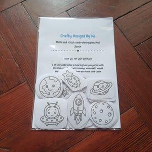 May include: A clear plastic bag containing white stick and stitch embroidery patches with black line art of space themes including a planet, rocket, and UFO. The included paper insert reads Crafty Designs By KJ, Stick and stitch embroidery patches, Space.