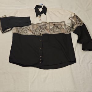 Op de afbeelding: Een overhemd met knopen, een zwarte body en een wit bovenstuk. Het bovenste gedeelte heeft een tijgerprint ontwerp. De mouwen zijn zwart met manchetten met knopen. Het shirt heeft een klassieke kraag en goudkleurige knopen.