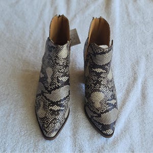 Puede incluir: Un par de botines con punta afilada y estampado de piel de serpiente en tonos negro, gris y crema. Los botines tienen un diseño con aberturas laterales, cierre de cremallera en la parte posterior y forro interior beige con etiqueta.