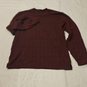 Könnte beinhalten: Ein langärmeliges, weinrotes Hemd mit gerippter Struktur. Das Hemd hat einen Rundhalsausschnitt und eine lockere Passform. Die Ärmel sind lang und enden mit einer Manschette. Das Hemd ist aus einem weichen, bequemen Material gefertigt.