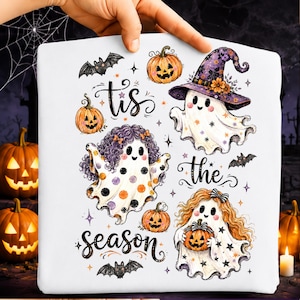 Puede incluir: Una camiseta blanca con un gráfico de Halloween que muestra tres fantasmas, calabazas, murciélagos y el texto 'tis the season'. Los fantasmas tienen peinados y accesorios distintos, incluyendo un sombrero de bruja morado, lunares y estrellas.