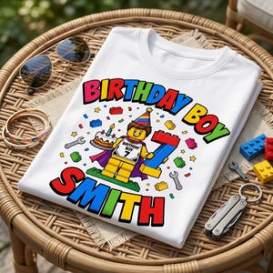 May include: A white t-shirt featuring a colourful graphic of a yellow toy figure wearing a party hat and cape, holding a birthday cake and a large number 7. The text reads BIRTHDAY BOY SMITH, surrounded by small toy bricks and tools.