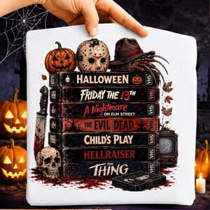 May include: A white t-shirt featuring a graphic of a stack of seven VHS tapes titled Halloween, Friday the 13th, A Nightmare on Elm Street, The Evil Dead, Child's Play, Hellraiser, and The Thing, with a pumpkin, hockey mask, and clawed glove.