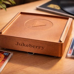 Può includere: Un lettore musicale Jukeberry in legno con un simbolo di pulsante di riproduzione sulla parte superiore. Il lettore è circondato da CD con le etichette "90s Hits" e "Rock Classics", e un disco in vinile. Il lettore ha la parola "Jukeberry" incisa.