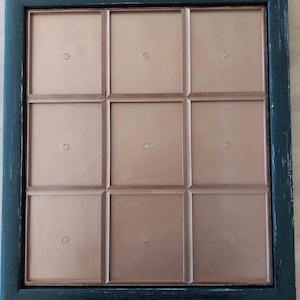 Puede incluir: Un marco de fotos de nueve secciones con un borde de madera negro desgastado. Cada sección tiene un interior de color cobre y un pequeño detalle circular en el centro, diseñado para contener fotos u obras de arte. El marco es cuadrado.