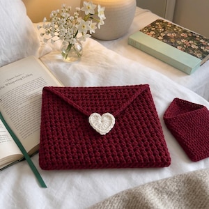 May include: A dark red crochet envelope-style pouch featuring a white heart-shaped button closure, accompanied by a smaller matching dark red crochet triangular pouch, both crafted with a textured stitch pattern on a white fabric surface.