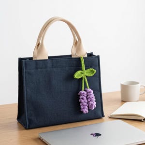 May include: A dark navy blue canvas tote bag with two beige fabric handles. A crocheted charm featuring two light purple flower clusters with green stems and leaves is attached to one handle by a small silver-toned metal ball chain.
