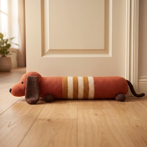 Peut inclure: Un butoir de porte en crochet en forme de teckel long et cylindrique, avec un corps orange rouille, des oreilles marron foncé, une petite queue et quatre minuscules pattes. Le centre du corps présente des rayures verticales crème, moutarde et marron.