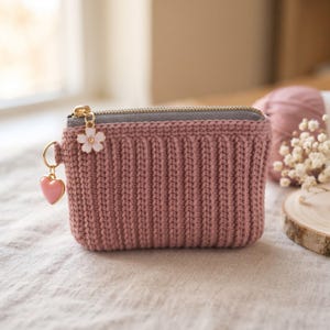 May include: A dusty rose crochet coin purse featuring a ribbed texture, a grey zip closure, and two gold-tone metal charms attached to the zip pull: one white flower with a gold centre and one small pink heart-shaped pendant.