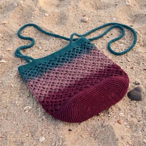 May include: A crochet shoulder bag featuring a gradient color pattern that transitions from deep teal at the top to a dusty rose in the middle and a solid dark burgundy at the base. The bag has an openwork mesh design and a long braided strap.
