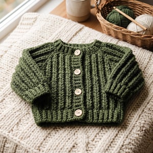 May include: A long-sleeved, olive green crochet cardigan featuring a vertical ribbed texture and four light-coloured wooden buttons down the centre front. The garment has a round neckline and a straight hem, showcasing a thick, chunky yarn construction.