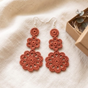 May include: A pair of rust-colored crochet earrings featuring a vertical arrangement of three floral motifs of varying sizes. Each earring is suspended from a silver-toned metal hook. The intricate thread work creates a delicate, openwork pattern.