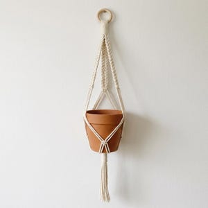 May include: A cream-colored macrame plant hanger featuring a circular wooden ring at the top, intricate braided cord patterns, and a central terracotta pot. The design concludes with a long, straight tassel hanging below the base of the pot.