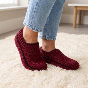 Puede incluir: Un par de zapatillas de ganchillo en color burdeos intenso con un patrón de punto texturizado y un fino borde negro a lo largo del cuello y el lateral. Las zapatillas son de corte bajo, cubren el pie hasta el tobillo y son de hilo grueso.