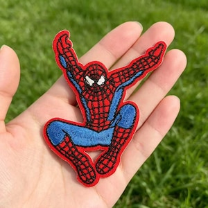 May include: An embroidered fabric patch featuring a superhero character in a dynamic crouching pose. The design displays a red and blue suit with a black web pattern, white eye lenses, and a thick red border outlining the entire figure shape.
