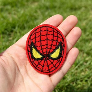 May include: A circular embroidered patch featuring a red background with a black web pattern and two large, almond-shaped yellow eyes outlined in black, designed to resemble the mask of a comic book character.