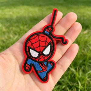 May include: A small embroidered patch featuring a stylized, chibi-style character with a red and blue suit, a webbed pattern on the head, and large white eyes. The character is depicted in a dynamic pose with a black line extending from the top.