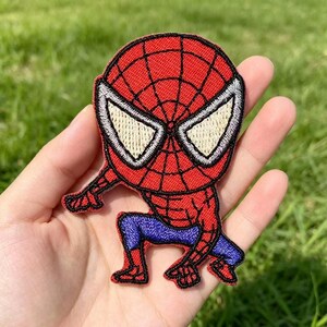 May include: An embroidered fabric patch featuring a stylised character with a red head and torso, a black web pattern, large white triangular eyes, and blue legs. The character is depicted in a crouching pose with one arm extended forward.