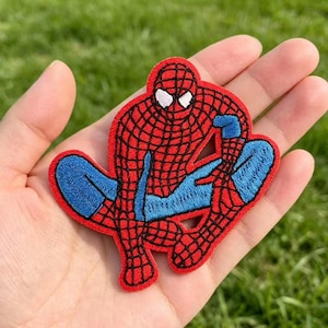 May include: An embroidered fabric patch featuring the superhero Spider-Man in a crouching pose. The design displays a red suit with a black web pattern, blue limbs, and white eye lenses. The patch has a distinct red border and textured stitching.