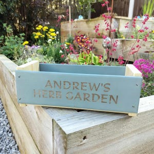 May include: A light blue wooden planter box with the text "ANDREW'S HERB GARDEN" in a brown font. The box is supported by wooden blocks and is set against a backdrop of a garden with flowers.
