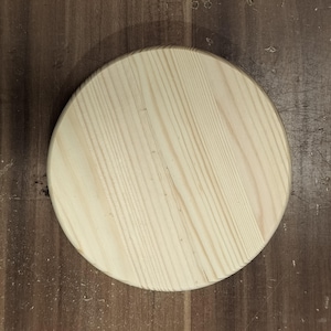 May include: A circular, unfinished light-coloured wooden plaque with a smooth surface and visible natural grain patterns running diagonally across the piece. The wood features pale cream and tan tones with darker linear grain details throughout.