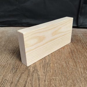 May include: A rectangular, unfinished wooden block with a light, natural wood grain. The block is angled on one side, creating a beveled edge. The wood has a light, warm tone, and the grain is visible.
