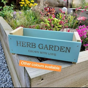 May include: A light blue wooden herb garden planter with the text "HERB GARDEN" and "GROWN WITH LOVE". The planter has natural wood corner supports and feet. The background shows a garden with flowers. The text "Other colours available" is also visible.