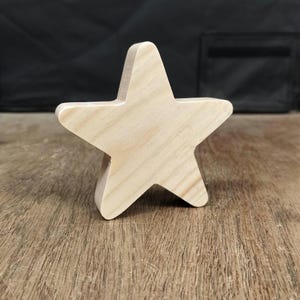 May include: A five-pointed star cutout made of light-coloured natural wood with a visible grain pattern. The star has smooth, flat edges and a thick profile, standing upright on its bottom point. The surface shows the raw texture of the wood.