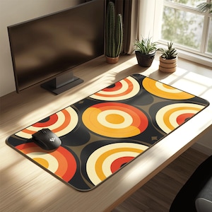 May include: A rectangular mouse mat with a retro design featuring concentric circles in orange, red, and cream colours on a black background. A black computer mouse is on the left side of the mat. The mat is on a wooden desk.